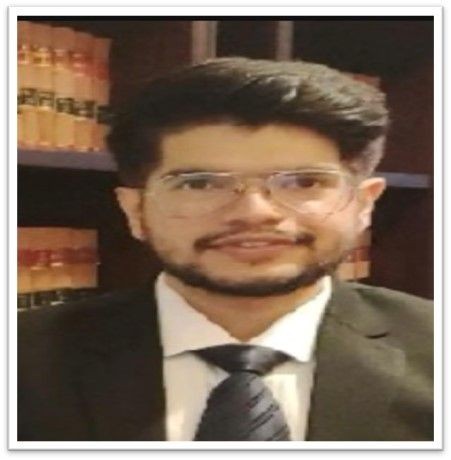 Individual wearing a formal suit and tie, standing in front of a bookshelf filled with legal books.